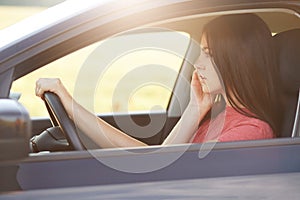 Horizontal shot of prosperous young businesswoman drives car, being in hurry for business meeting, has telephone conversation with