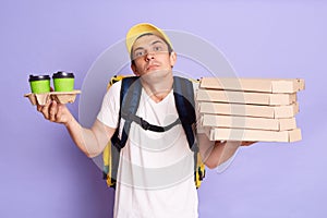 Horizontal shot of puzzled man in yellow cap and t-shirt, holding pizza and paper cups of coffee isolated on lilac background,