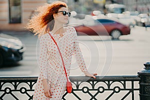 Horizontal shot of red haired woman wears sunglasses, focused aside, poses near hence at street, poses against road with transport