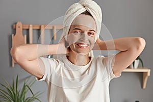 Horizontal shot of relaxed Caucasian woman with clay mask for face skin and being wrapped in towel posing in kitchen, raised her