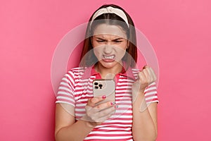 Horizontal shot of sad irritated young girl with hair band wearing striped casual t shirt, holding with smart phone in hands,