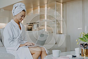 Horizontal shot of satisfied beautiful woman dressed in white soft bathrobe works on laptop computer surfes internet poses near