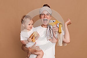 Horizontal shot of satisfied joyful woman with dark hair wearing white t shirt standing with her baby daughter and screaming with