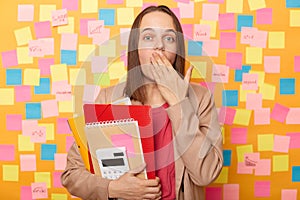 Horizontal shot of shocked astonished woman with brown hair wearing beige jacket, covering mouth with palm, forget something