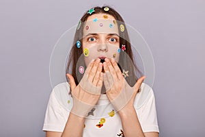 Horizontal shot of shocked astonished woman covered with funny stickers posing isolated over gray background, covering mouth, sees