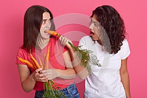 Horizontal shot of shocked emotional female having carrot in mouth, eating vegetables. Charismatic brunette giving vegetables for