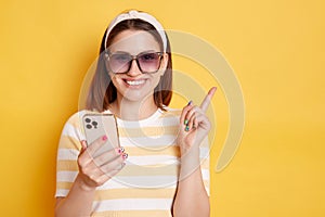 Horizontal shot of smiling happy young woman wearing striped t-shirt posing isolated over yellow background, holding cell phone
