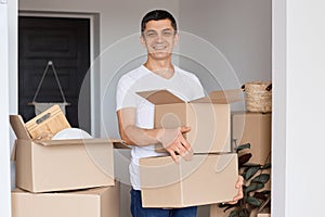 Horizontal shot of smiling man wearing white t shirt standing with cardboard box in hands, looking at camera with smile, being