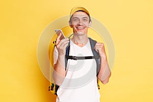 Horizontal shot of smiling optimistic delivery man in yellow cap standing with thermal backpack holding mobile phone, using gadget