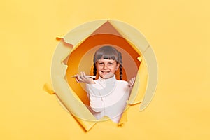 Horizontal shot of smiling satisfied little girl with pigtail breaking through yellow paper and holding mobile phone, using voice