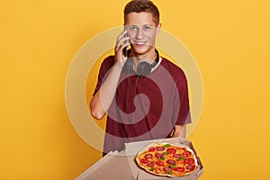 Horizontal shot of smiling sincere delivery man talking over phone, having headphones around neck, delivering one pizza cardbox,