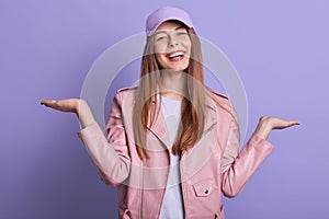 Horizontal shot of smiling teen girl with spreading hands aside, expressing happyness, posing isolated over lilac background,