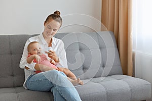 Horizontal shot of smiling woman wearing white shirt and jeans sitting on sofa with baby daughter, cute kid drinking water from