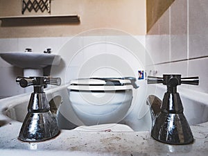 Horizontal shot of a steel bath taps on a background of a toilet seat in a dirty bath and sink