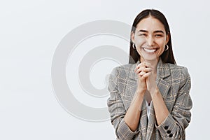 Horizontal shot of stylish happy european girlfriend in jacket, clasping hands together over chest and smiling joyfully