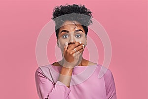 Horizontal shot of surprised African American woman covers mouth with both hands, tries to be speechless, looks with