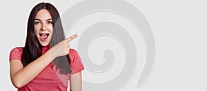 Horizontal shot of surprised brunette woman with dark hair, dressed in pink t shirt, points with index finger asie, shows free spa