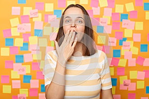 Horizontal shot of surprised Caucasian woman wearing striped t shirt, sees something astonishing, saying wow, covering mouth with