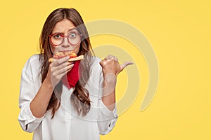 Horizontal shot of surprised female eats tasty slice of pizza, dressed in fashionable clothes, indicates with thumb