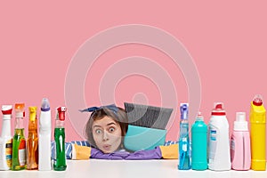 Horizontal shot of surprised housemaid looks in stupor, leans at hands on table, carries broom, surrounded with many