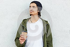 Horizontal shot of thoughtful dreamy teenage boy in stylish outfit, drinks hot aromatic coffee or cappuccino, looks pensively asid photo