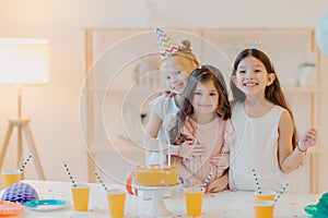 Horizontal shot of three happy friends embrace and have funny expressions, pose near festive table with cake indoor. Three girls