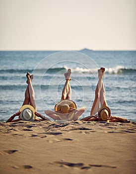 Horizontal shot of three young women in straw hats on a beach, with legs up. Blue sea in the background. Summer vacation concept