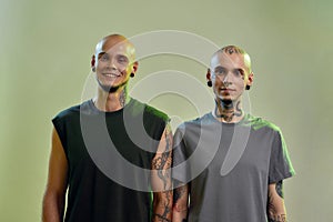 Horizontal shot of two happy young twin brothers with tattoos and piercings smiling at camera while posing together in