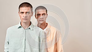 Horizontal shot of two young caucasian men, twin brothers looking at camera while posing together isolated over beige