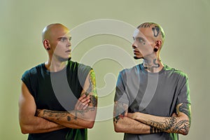 Horizontal shot of two young caucasian twin brothers with tattoos and piercings keeping arms crossed and looking at each