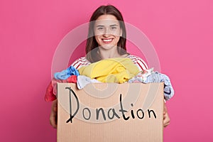 Horizontal shot of volunteer female standing isolated over pink background in studio, holding box with inscription donation,