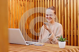 Horizontal shot of winsome smiling woman with bun hairstyle holding smart phone and recording voice message, female wearing beige