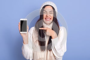 Horizontal shot of woman showing smart phone with blank screen and pointing at It, posing  over blue background, winsome