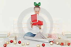 Horizontal shot of woman sitting on bed at home, in cosy bedroom, wearing casual clothes, preparing xmas presents, wrapping gift