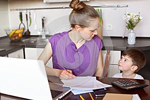 Horizontal shot of working mum being busy with documentation, makes financial report or calculates family budget, surrounded with photo