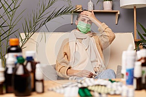 Horizontal shot young adult woman wearing warm sweater, scarf and protective mask sitting on sofa in home interior among pills,