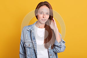 Horizontal shot of young charming woman in denim jacket and white casual shirt standing over yellow background, touches her