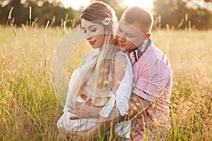 Horizontal shot of young family embrace and keep hands on tummy, anticipate for child, pose in green field, have walk during sunny