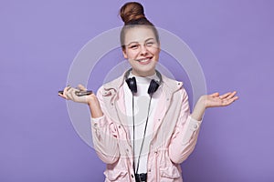 Horizontal shot of young pretty girl with hairbun listening to music and spreading palms aside, having headphones around neck,