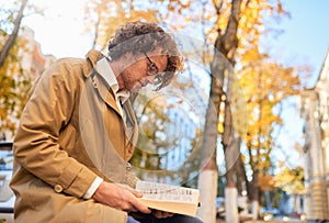 Horizontal side view image of trendy handsome young man reading book outdoors. College male student carrying books in campus in