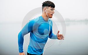 Horizontal side view image of young athelte man running in the morning on the fog lake background. Fitness male exercising in the