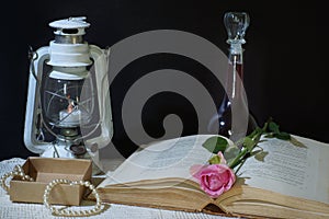 Horizontal still life on a black background with a book, an old lantern and a bottle of wine, a pink rose lies between the pages,