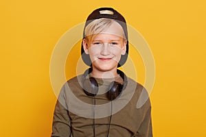 Horizontal studio picture of blond haired sweet kid having pleasant facial expression, smiling sincerely, having headphones around