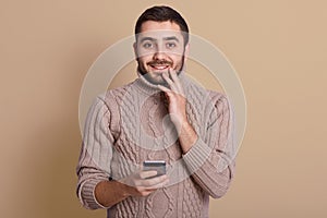 Horizontal studio picture of pleasant delighted young man looking directly at camera, touching chin with hand, holding his