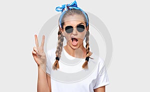 Horizontal studio portrait of cheerful blonde young woman wearing trendy sunglasses, white t-shirt and blue headband, showing