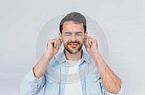 Horizontal studio portrait of handsome bearded young male plugs ears with displeased expression as hears annoying loud noise,