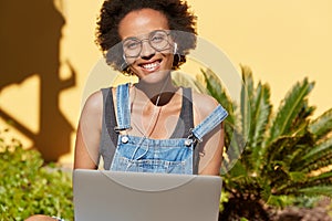 Horizontal view of black youngster with toothy smile, enjoys blogging, works on laptop computer, listens audio lesson