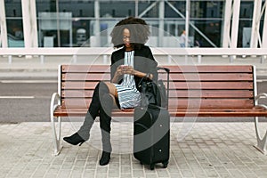 Horizontal view of the joyful afro-american woman is sitting on the bench and texting via the cellphone near the airport