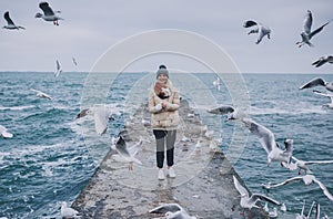 Horizontal view of young happy woman feeds seagulls on the sea. Pretty female wearing coat, scarf and watching flying seagulls by