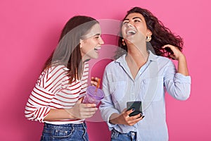 Horizontalshot of pretty young women laughing happily, have phone and water bottle in hands, wearing stylish clothing, posing photo